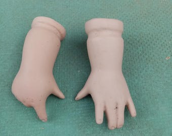 Może przedstawiać: Dwie antyczne porcelanowe dłonie lalki, w kolorze bladego różu, z szczegółowymi palcami i nadgarstkami. Dłonie są oddzielone i wydają się pochodzić ze starej lalki, prezentując delikatną i postarzaną estetykę.