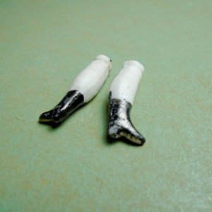 May include: Two antique doll legs. The legs are white porcelain with black painted boots. The boots have a glossy finish. The legs are resting on a light green surface.