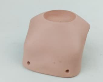 May include: A light pink, hollow mannequin torso with a rounded neck and two small holes at the bottom. The form is smooth and appears to be made of a solid material, likely plastic or ceramic. The design is simple and functional.