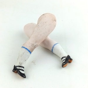 May include: A pair of porcelain doll legs, crossed at the ankles. The legs are painted with a pale skin tone, white socks with blue trim, and black and white striped shoes with small heels.