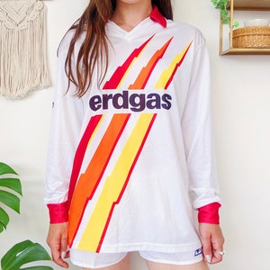 May include: White long-sleeve jersey with a red, orange, and yellow lightning bolt design. The jersey has a black "erdgas" logo on the front.
