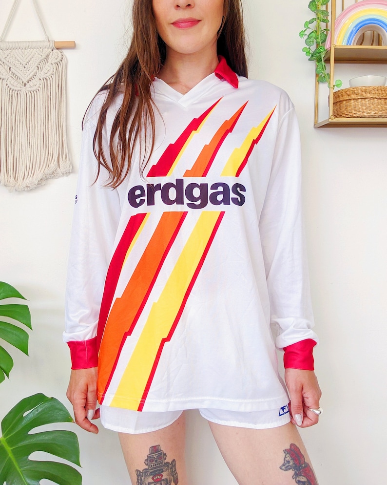 May include: White long-sleeve jersey with a red, orange, and yellow lightning bolt design. The jersey has a black "erdgas" logo on the front.