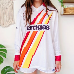 May include: White long-sleeved soccer jersey with a red, orange, and yellow lightning bolt design and the word "erdgas" in black letters.