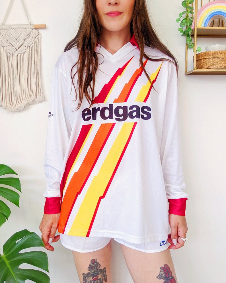 May include: White long-sleeved soccer jersey with a red, orange, and yellow lightning bolt design and the word "erdgas" in black letters.