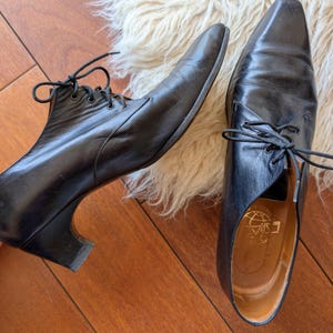 Peut inclure: Une paire de bottines en cuir noir à lacets avec des bouts pointus. Une botte a un petit talon. Les bottes sont sur une surface en bois avec un tapis blanc et moelleux en arrière-plan. L'intérieur d'une botte porte des lettres dorées.