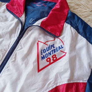 May include: A white, blue and red windbreaker jacket with a zip closure. The jacket has a large graphic on the front that says "ÉQUIPE MONTRÉAL 98".
