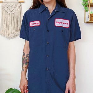 Op de afbeelding: Een donkerblauw werkshirt met korte mouwen en een knoopsluiting. Het shirt heeft twee opgestikte borstzakken, elk met een rood en wit naamschild. De linkerzak heeft de naam "Emory" en de rechterzak heeft de naam "Roger Wood".