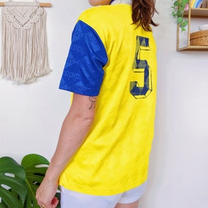 May include: Yellow soccer jersey with blue sleeves and a blue number 5 on the back. The sleeves have a repeating pattern of blue squares with white diagonal lines.
