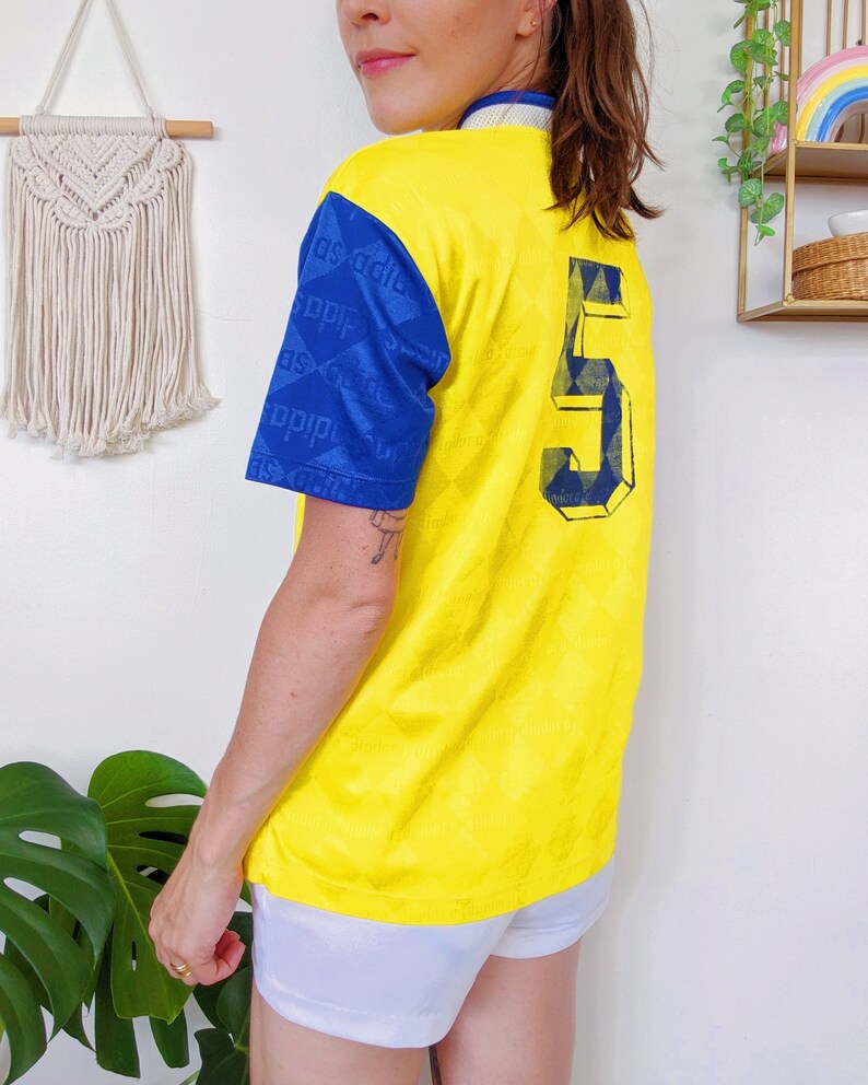 May include: Yellow soccer jersey with blue sleeves and a blue number 5 on the back. The sleeves have a repeating pattern of blue squares with white diagonal lines.