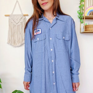 May include: A light blue long-sleeved work shirt with a patch that says "CDK", a white collar, and two chest pockets.