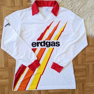 May include: White long-sleeve soccer jersey with red and yellow stripes and the word "erdgas" printed on the front.
