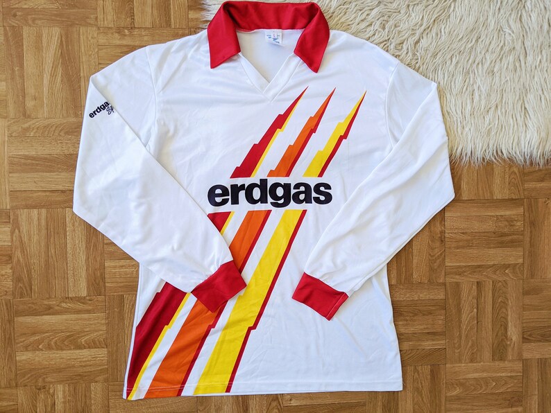 May include: White long-sleeve soccer jersey with red and yellow stripes and the word "erdgas" printed on the front.