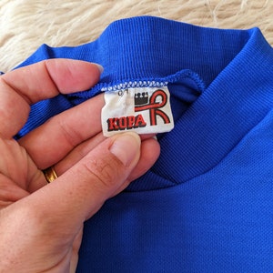 May include: A close-up of a blue shirt collar with a white tag that reads "KOPA" in red letters with a red ribbon and crown logo.