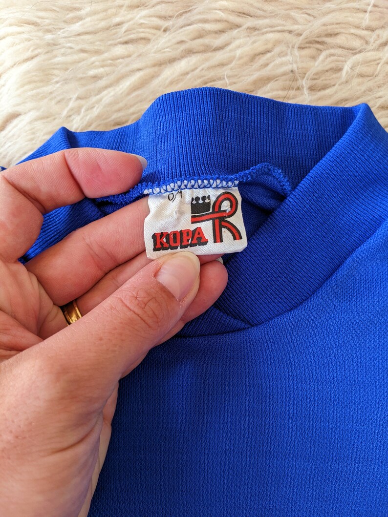 May include: A close-up of a blue shirt collar with a white tag that reads "KOPA" in red letters with a red ribbon and crown logo.