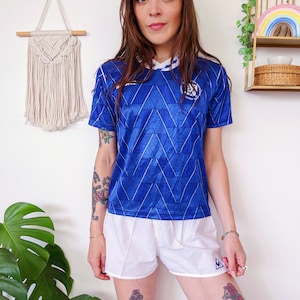 Puede incluir: Una camiseta de fútbol azul y blanca con un patrón geométrico. La camiseta tiene un cuello blanco con una franja azul y un logotipo blanco con el texto "A.S.O. 1950" en la parte delantera.