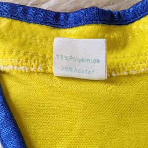 May include: Close-up of a white fabric label sewn into a yellow garment. The label reads "72% Polyamide 28% Acetat."