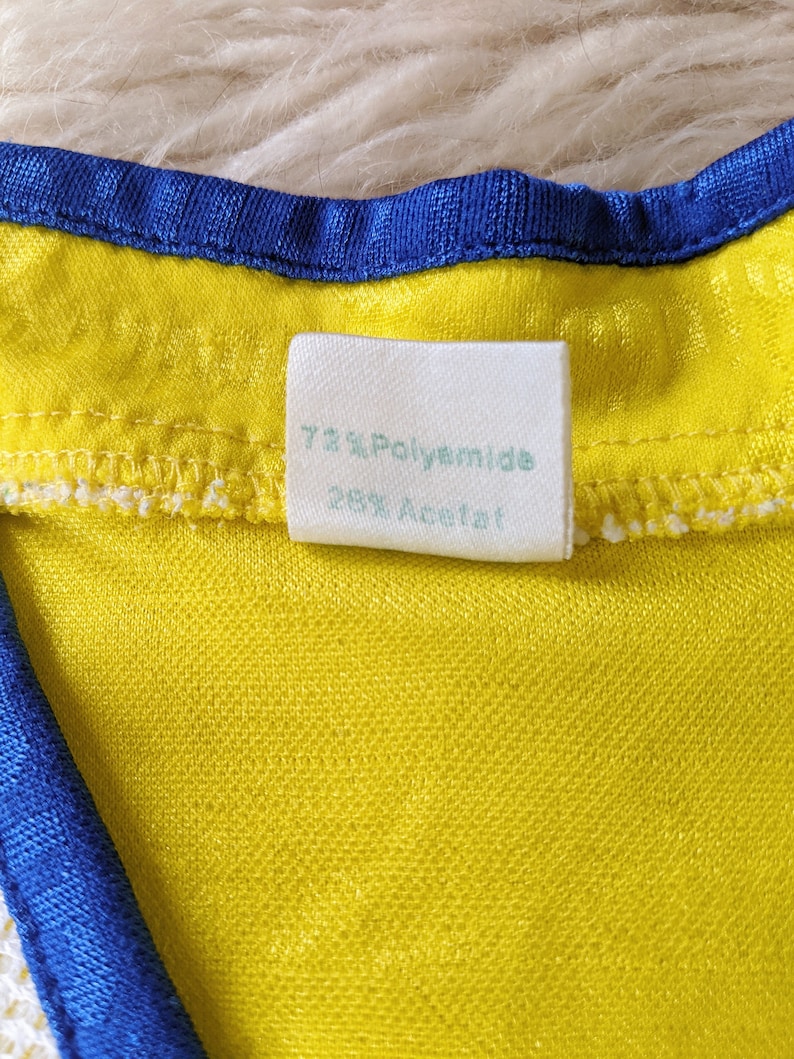 May include: Close-up of a white fabric label sewn into a yellow garment. The label reads "72% Polyamide 28% Acetat."