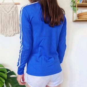 May include: A royal blue long-sleeved shirt with white stripes down the sleeves. The shirt has a high neckline and a relaxed fit.