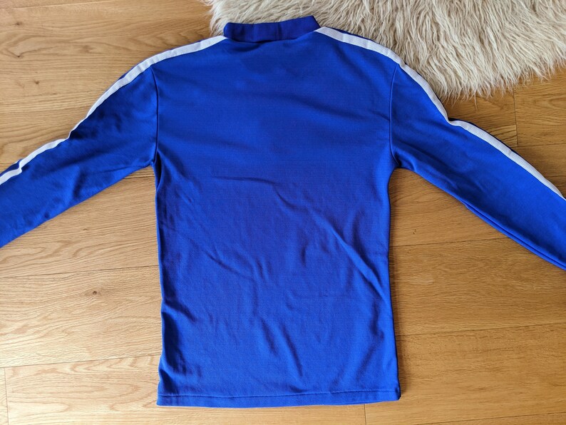 May include: A long-sleeved blue shirt with white stripes down the sleeves. The shirt has a high neck.