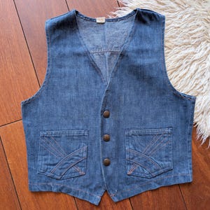 以下が含まれることがあります： Vネック、2つのフロントポケット、3つの茶色のボタンが付いた、ヴィンテージの青いデニムベスト。ポケットには装飾的なステッチが施されています。ベストはおそらくSまたはMサイズです。