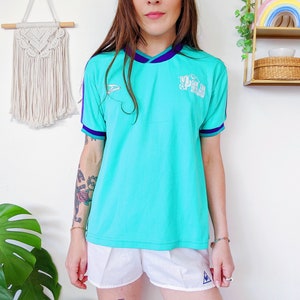 Peut inclure: Un t-shirt turquoise à manches courtes avec un logo blanc qui dit "San Tan Soccer Association". Le t-shirt a une bordure violette autour du cou et des manches. Le mannequin porte un short blanc avec un logo triangulaire bleu.