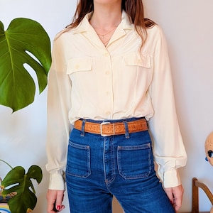 Puede incluir: Una camisa crema con botones y dos bolsillos en el pecho, combinada con vaqueros azules de cintura alta y lavado oscuro. Un cinturón marrón con diseño adorna la cintura. El conjunto tiene una estética vintage.