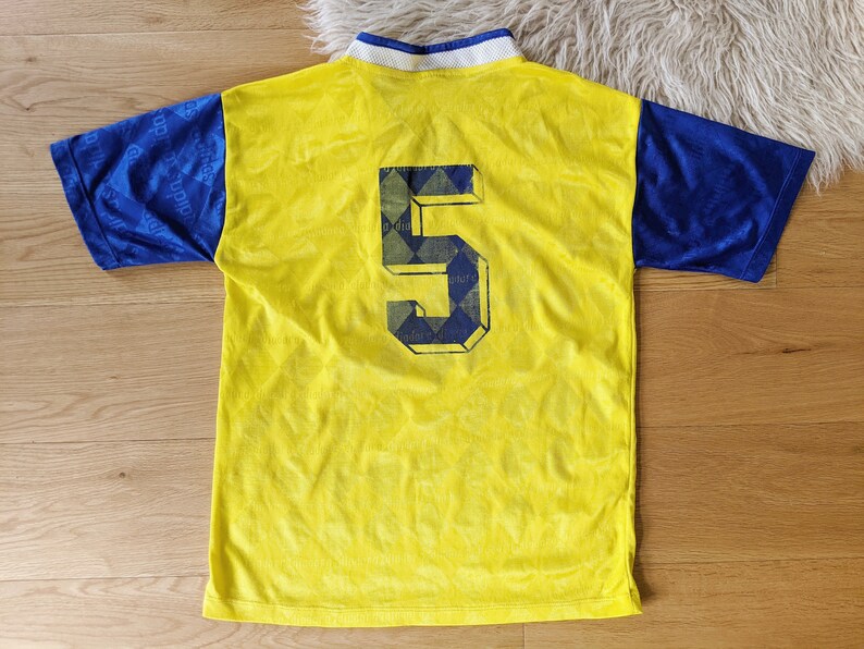 May include: Yellow and blue soccer jersey with the number 5 on the back. The jersey has a diamond pattern on the back.