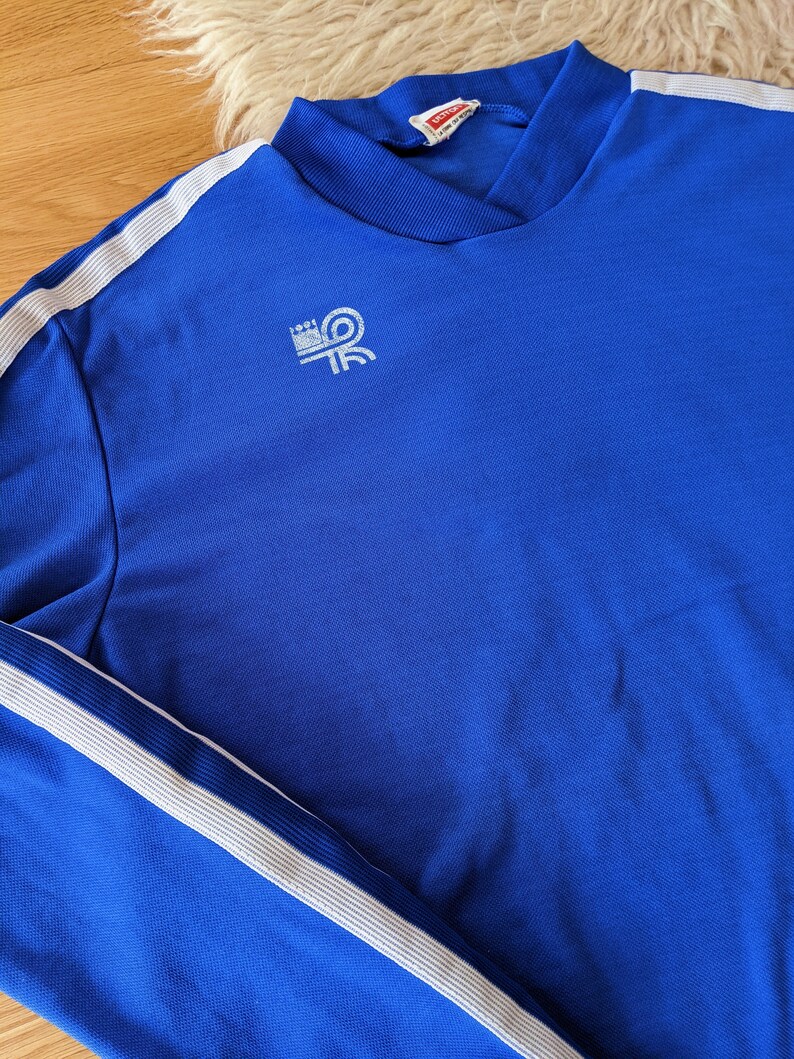 May include: A royal blue long-sleeve shirt with white stripes down the sleeves and a white logo on the left chest that says "LOUI" with a stylized "R" inside a circle.