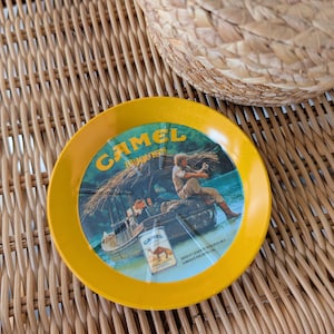 Op de afbeelding: Een gele asbak met een Camel-sigarettenreclame met een man op een boot in een tropische omgeving. De tekst op de asbak luidt "Camel  Turkish & Domestic Blend  R.J. Reynolds Tobacco Co. Winston-Salem, N.C."