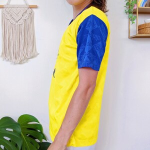 May include: Yellow short-sleeved t-shirt with blue and white patterned sleeves. The sleeves have the text "adidas" repeated in a pattern.