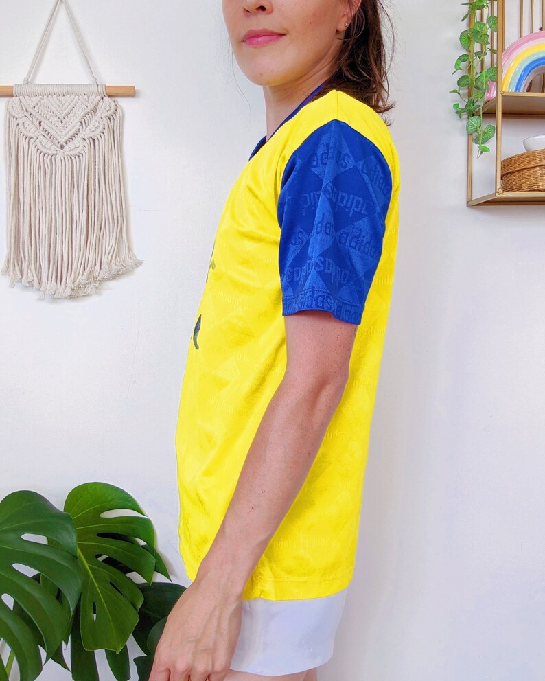 May include: Yellow short-sleeved t-shirt with blue and white patterned sleeves. The sleeves have the text "adidas" repeated in a pattern.