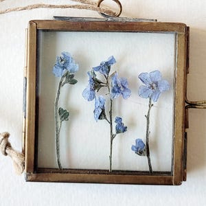 May include: A square, vintage-style brass frame with a glass front, showcasing delicate blue dried flowers. The frame has a small loop at the top for hanging and is suspended by a natural twine cord. The flowers are arranged against a white background.
