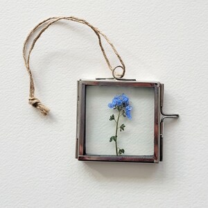 May include: A small silver frame with a blue flower pressed inside. The frame has a string attached to it, making it a hanging ornament.