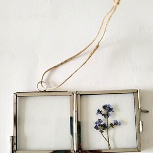 May include: A silver metal picture frame with a hinged design. The frame is hanging from a twine string and contains a pressed flower with blue petals.