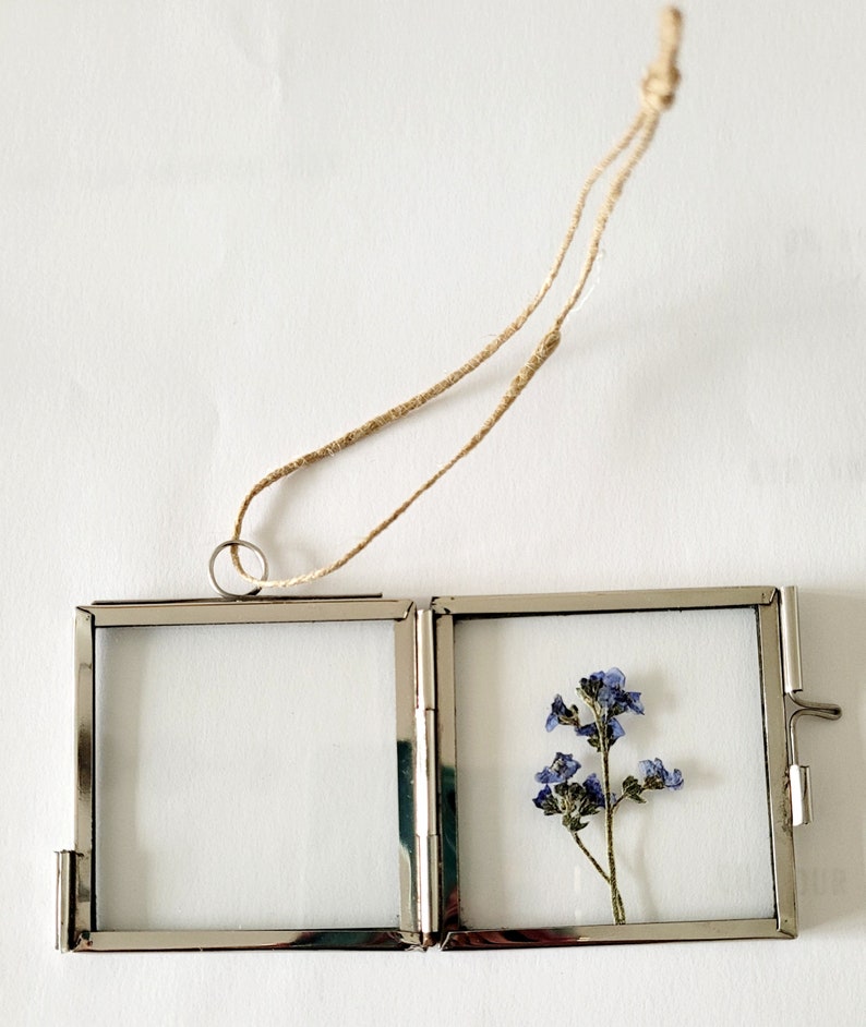 May include: A silver metal picture frame with a hinged design. The frame is hanging from a twine string and contains a pressed flower with blue petals.