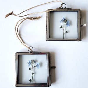 May include: Two small silver picture frames with a delicate blue flower pressed inside each frame. The frames have a string attached for hanging.