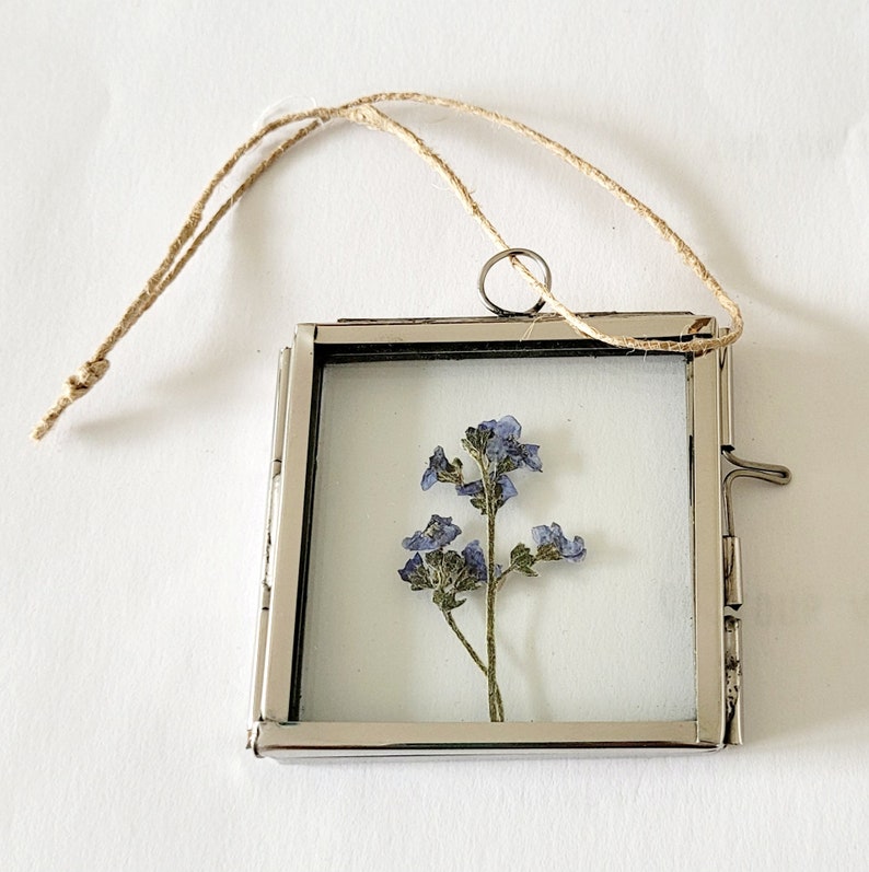 May include: A silver metal frame with a clear glass insert, featuring a pressed flower arrangement of blue forget-me-nots. The frame has a hanging loop and a small latch on the side.
