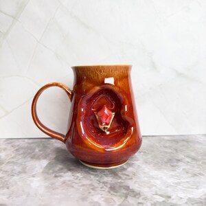 May include: A red and brown ceramic mug with a unique, textured design. The mug has a handle and a small, red, gemstone-like detail on the front.
