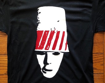 May include: Black t-shirt with a white and red graphic of a person wearing a bucket hat. The hat has red stripes and the person's face is white.