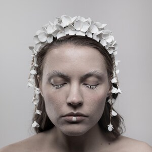 May include: A woman wearing a white flower crown with gold chain accents. The crown is made of delicate white flowers and has a simple, elegant design. The woman has her eyes closed and is wearing white makeup.