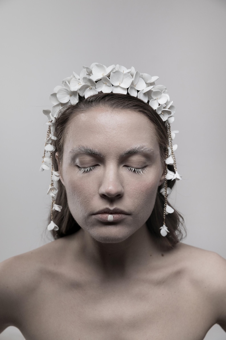 May include: A woman wearing a white flower crown with gold chain accents. The crown is made of delicate white flowers and has a simple, elegant design. The woman has her eyes closed and is wearing white makeup.