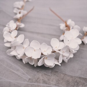 May include: A white floral crown with delicate white flowers and a gold wire base. The crown is lying on a white tulle fabric.