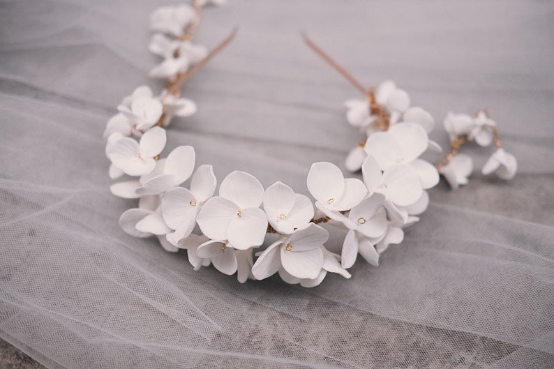 May include: A white floral crown with delicate white flowers and a gold wire base. The crown is lying on a white tulle fabric.