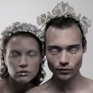 May include: Two people with their eyes closed wearing white flower crowns. The crowns are made of delicate white flowers and have a pearl accent. The people are wearing white makeup and have white accents on their eyelashes.