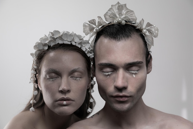 May include: Two people with their eyes closed wearing white flower crowns. The crowns are made of delicate white flowers and have a pearl accent. The people are wearing white makeup and have white accents on their eyelashes.