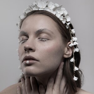 May include: A woman with her eyes closed wears a white flower crown with a gold chain and white flower accents. The crown is made of delicate white flowers and has a gold chain that hangs down the side of her face.