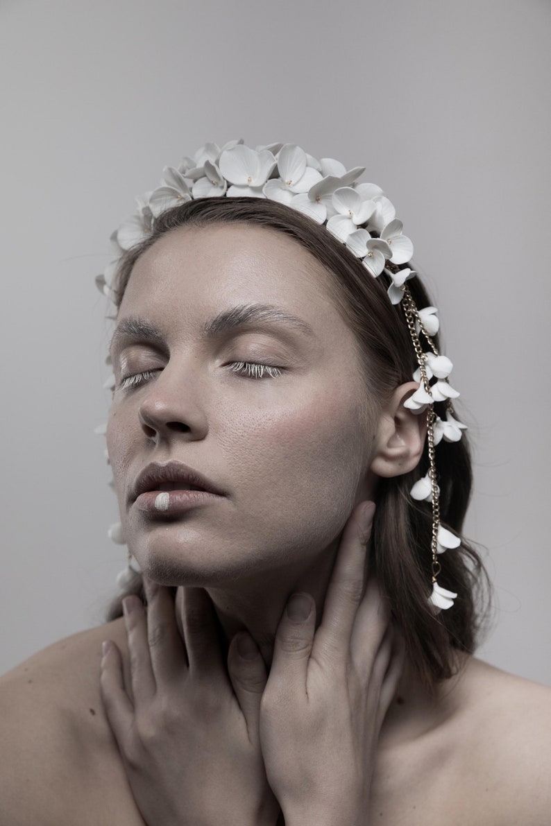 May include: A woman with her eyes closed wears a white flower crown with a gold chain and white flower accents. The crown is made of delicate white flowers and has a gold chain that hangs down the side of her face.
