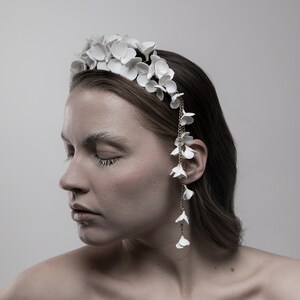 May include: A white floral headband with a delicate chain and white flower accents. The headband is worn by a person with brown hair.