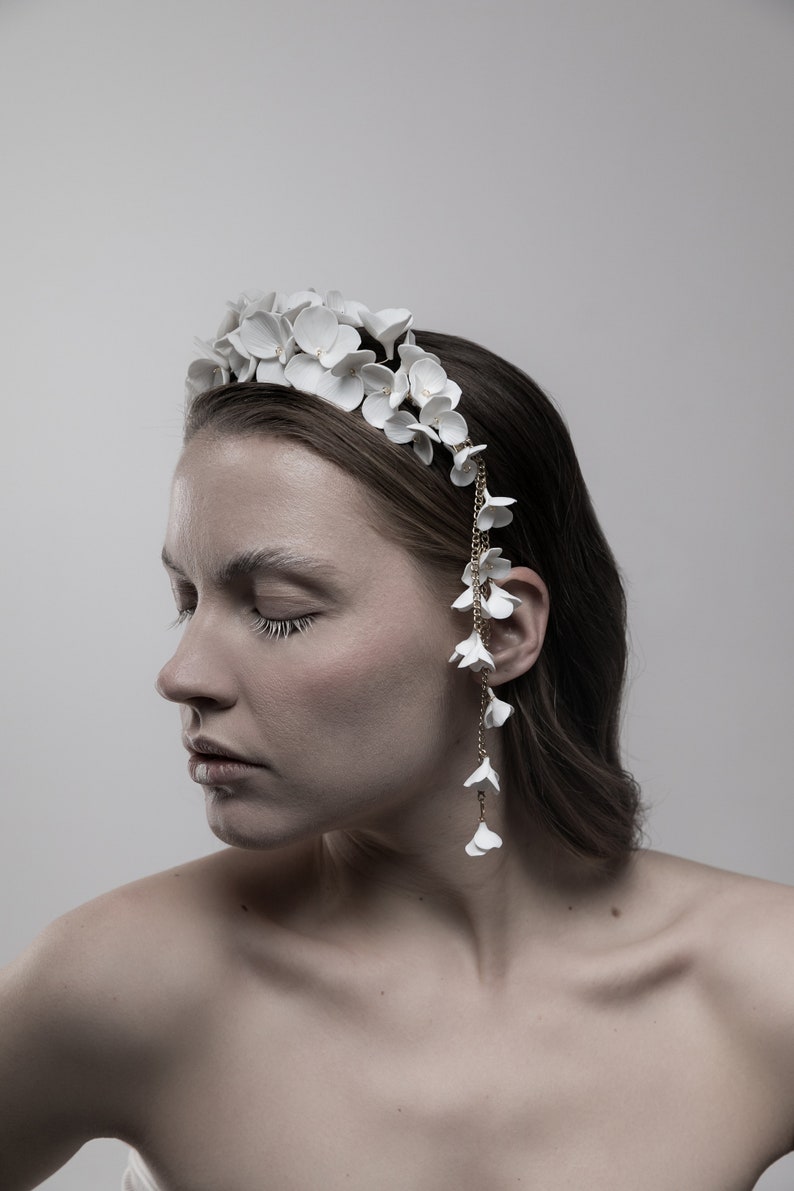 May include: A white floral headband with a delicate chain and white flower accents. The headband is worn by a person with brown hair.