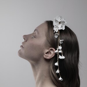 May include: A white floral headband with a gold chain and white flower accents. The headband is worn by a person with long brown hair.